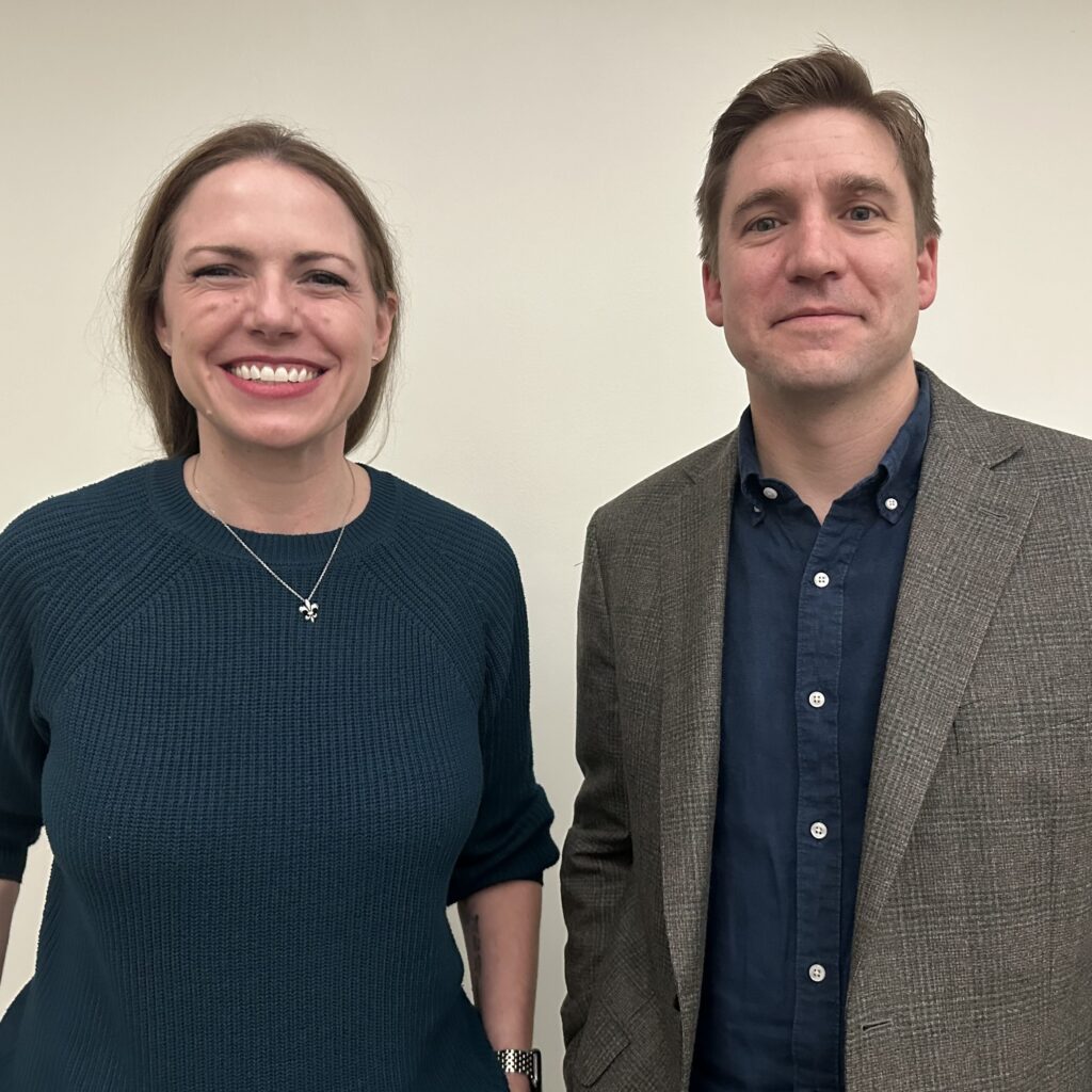 Carolyn Scofield (left) and Ryan Lauth (right) standing and smiling in front of a blank wall
